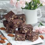 Low/no sugar chocolate nut bars with oats and pecans, arranged beside soft pink flowers.